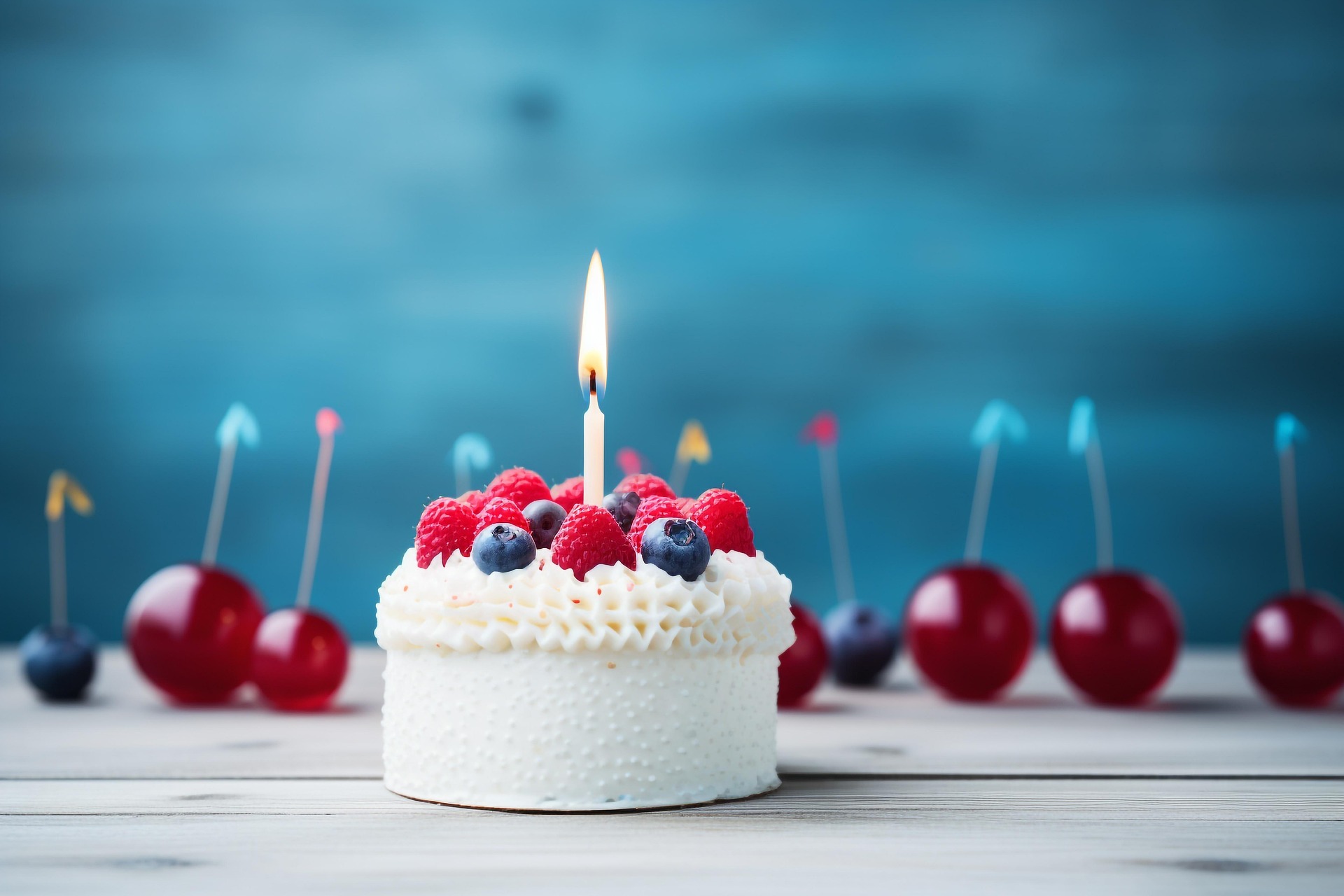Photographie d'un petit gâteau à la crème et aux fruits rouges, portant une bougie allumée. Il y a des fruits
(cerises, myrtilles) et un fond bleu en arrière plan.
